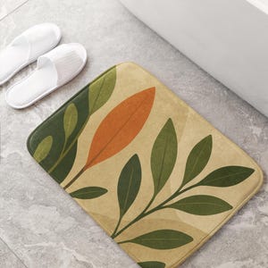 Peut inclure: Tapis de bain rectangulaire avec un motif botanique de feuilles vertes et orange sur fond beige. Le tapis est posé sur un sol carrelé gris, avec des pantoufles blanches et une baignoire blanche en arrière-plan.