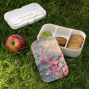 May include: A white lunch box with a floral design lid, containing grapes, cookies, and crackers, sits open on green grass. A red apple and a separate lid with a fork and spoon are also visible. The lunch box is divided into three compartments.