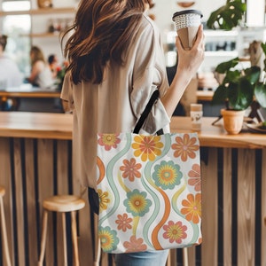 May include: A white tote bag with a retro floral pattern. The design features orange, yellow, pink, and blue flowers with wavy green and orange lines. The bag has a black strap.