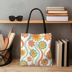 May include: A white tote bag with a colorful floral pattern in orange, yellow, and teal. The design features stylized flowers and wavy lines. The bag has black handles and is displayed on a wooden shelf.