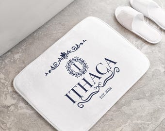 Puede incluir: Alfombra de baño blanca con un monograma azul marino y el texto "ITHACA EST. 2024".