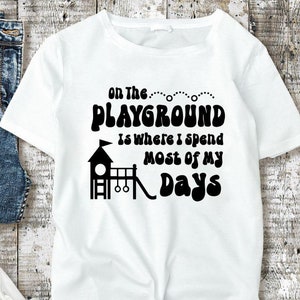 May include: White t-shirt with black text that reads "On the Playground is where I spend most of my days". The text is in a retro font and includes a black silhouette of a playground with a slide, swings, and a climbing structure.