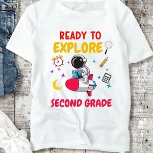 May include: White t-shirt with the text "READY TO EXPLORE SECOND GRADE" and a graphic of an astronaut on a rocket ship. The image also includes blue jeans and white sneakers on a wooden surface.