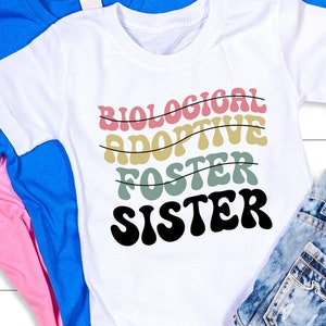 Op de afbeelding: Wit T-shirt met de tekst "BIOLOGICAL ADOPTIVE FOSTER SISTER" in een retro lettertype met gekleurde strepen. De tekst is in zwart, roze, geel en groen.