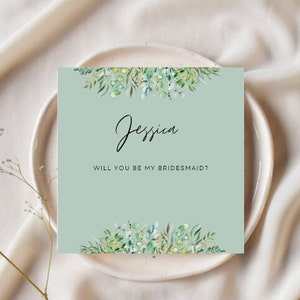 May include: A light green card with a floral design at the top and bottom. The card says "Jessica, will you be my bridesmaid?" in black text.