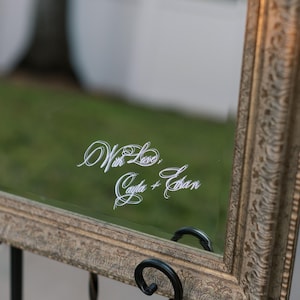 Puede incluir: Espejo decorativo con marco dorado ornamentado, sobre un soporte de metal negro. El espejo refleja un fondo verde y presenta el texto "With Love, Cayla + Ethan" en elegante escritura blanca.
