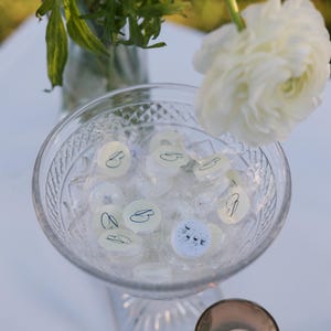 May include: A clear glass bowl filled with wrapped candies, some with a 'B' design, sits on a white surface. A lit candle in a small glass holder is next to the bowl. White flowers and greenery are in the background.