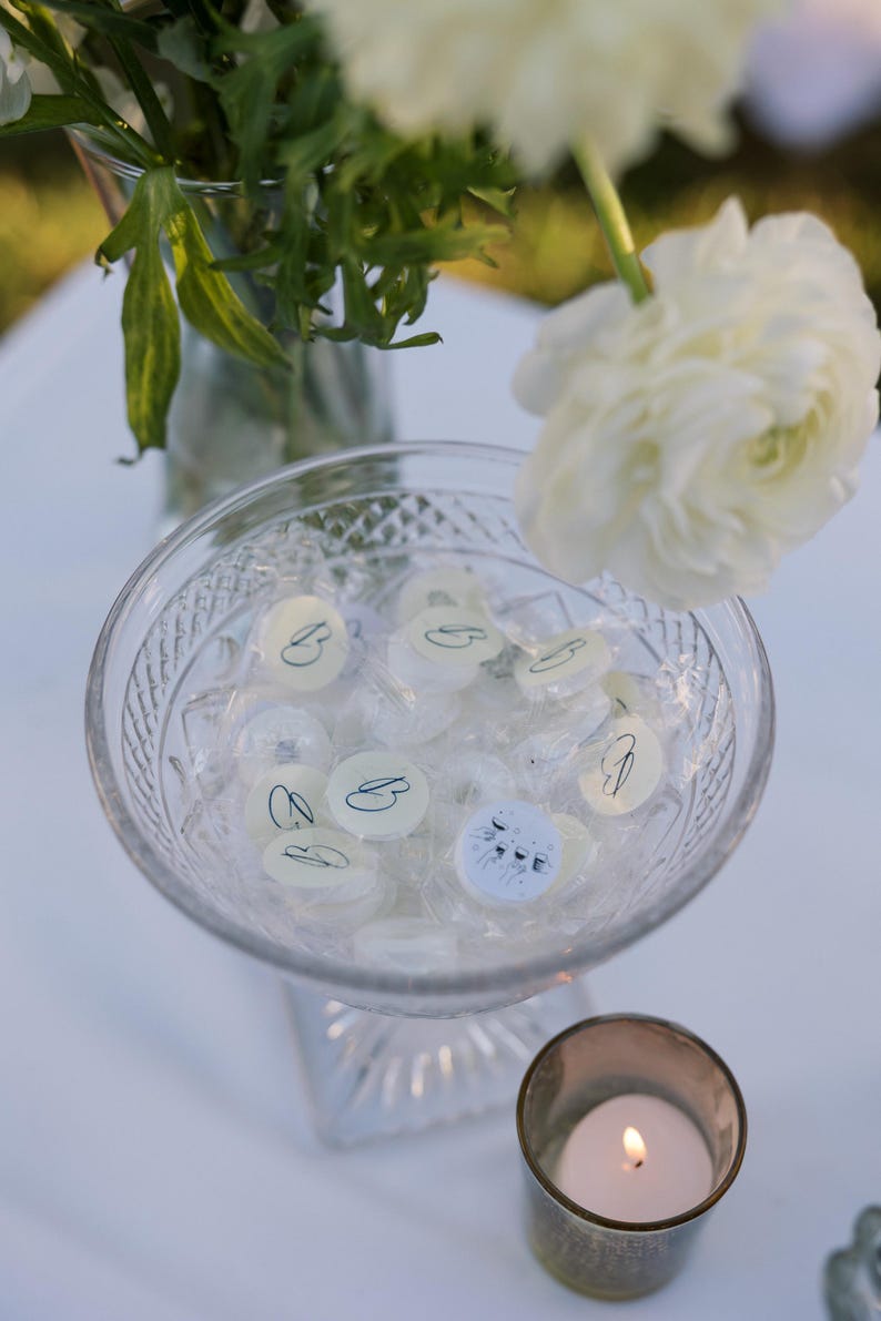 May include: A clear glass bowl filled with wrapped candies, some with a 'B' design, sits on a white surface. A lit candle in a small glass holder is next to the bowl. White flowers and greenery are in the background.