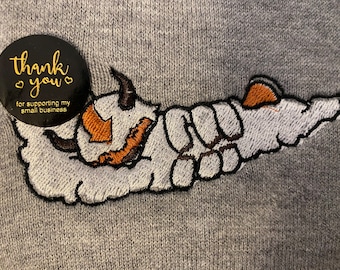 Può includere: Felpa grigia con un'immagine ricamata di una creatura bianca con dettagli arancioni e neri. La creatura è sdraiata con le braccia distese. Un adesivo nero e dorato con il testo "thank you for supporting my small business" è attaccato alla felpa.