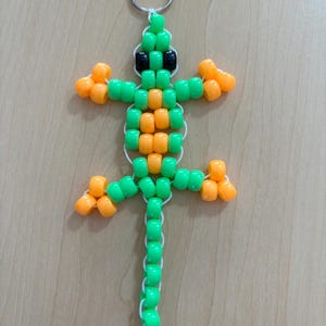 Puede incluir: Un llavero de lagarto hecho con cuentas verdes, naranjas y negras. El lagarto está hecho de cuentas de colores ensartadas con hilo blanco y tiene un llavero plateado en la parte superior. El cuerpo del lagarto es verde y naranja, con cuentas negras para los ojos.