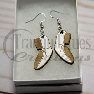 May include: A pair of wooden earrings shaped like white shoes with brown accents. The earrings are hanging from silver hooks.