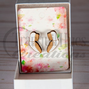 May include: Wooden ballet shoe earrings displayed in a white gift box. The earrings are white and brown with a delicate string detail. The background features a floral design with pink and white blossoms. The box is open, revealing the earrings.