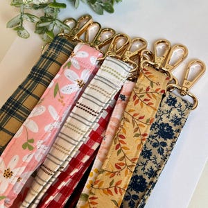 May include: An assortment of fabric lanyards with gold-coloured swivel clasps. The lanyards display diverse patterns, such as floral, plaid, and leaf motifs. The colour scheme includes pink, red, yellow, and blue.
