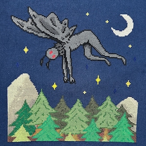 May include: A cross-stitch embroidery on dark blue fabric depicting a winged creature with red eyes flying over a forest of green pine trees. The night scene includes a white crescent moon and several yellow four-pointed stars in the sky.