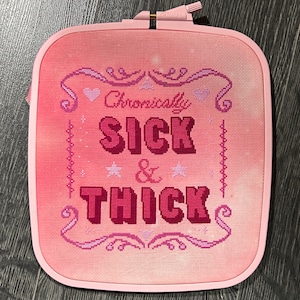 Pode incluir: Um bordado de ponto cruz rosa num bastidor quadrado. O texto "Chronically SICK & THICK" é bordado em tons de rosa e vermelho, com elementos decorativos como corações e estrelas. O bastidor é de cor rosa claro.