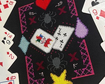 May include: A black cross-stitch design with playing card motifs, spiders, and a heart. The central design includes two playing cards, a red heart, and a yellow star. Playing cards surround the design.