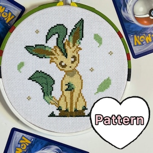 May include: A cross-stitch pattern of a green and yellow Pokemon character, Leafeon, on a white background. The pattern is framed in a white embroidery hoop with a green and red border. The word "Pattern" is written in a pink heart shape.