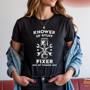 Puede incluir: Camiseta negra con el texto blanco "Knower of Stuff Fixer of Things". El diseño incluye un pistón, llaves y estrellas. La camiseta es de cuello redondo y la lleva una persona.