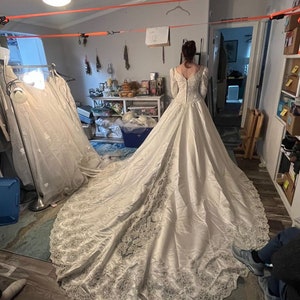 May include: A white satin wedding dress with a long train and lace detailing. The dress has a fitted bodice and long sleeves.