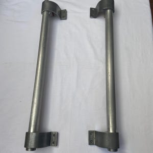 Può includere: Due barre di supporto in metallo con finitura argentata. Ogni barra ha un corpo cilindrico ed è collegata a una staffa curva ad ogni estremità. Le staffe hanno fori di montaggio. Le barre sono probabilmente per mobili o costruzioni.