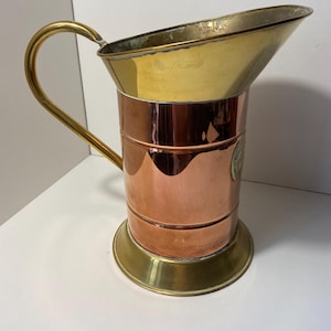 Könnte beinhalten: Eine Kupfer- und Messingkanne mit gebogenem Griff. Ober- und Unterteil sind aus Messing, während der Körper aus Kupfer besteht. Die Kanne hat einen Ausguss und ein dekoratives Emblem an der Seite. Die Kanne ist wahrscheinlich Vintage.