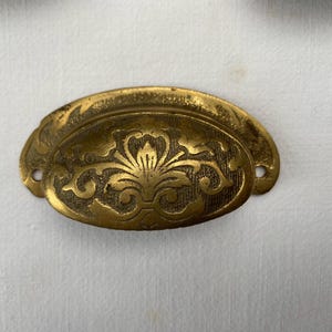 Peut inclure: Une poignée de tiroir en laiton de style vintage avec un motif floral orné. La poignée est de forme ovale avec une poignée incurvée et deux trous de vis. Le laiton a un aspect vieilli et antique.