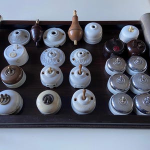 Pode incluir: Uma coleção de interruptores de luz e luminárias vintage numa bandeja de madeira escura. Os interruptores variam em estilo, incluindo porcelana branca, baquelite castanha e acabamentos cromados. Alguns têm alavancas de alternância, outros designs de botões. Também existem luminárias de teto decorativas.