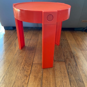 Può includere: Un tavolino da salotto rotondo di colore arancione brillante con un design unico. Il tavolino ha un piano circolare e quattro gambe che sono collegate al piano da un elemento di design unico. Il tavolino è in plastica e ha una finitura lucida.