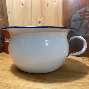 Può includere: Pentola da camera in smalto bianco con un bordo blu e un manico. La pentola è appoggiata su una superficie di legno.