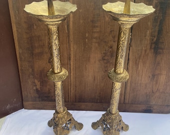 Puede incluir: Dos candelabros antiguos de color dorado. Cada uno tiene un plato ancho y festoneado en la parte superior, un tallo largo y ornamentado y una base decorativa. Los candelabros son de metal y tienen un aspecto vintage.