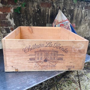 Peut inclure: Caisse à vin rectangulaire en bois clair avec l'inscription "Château La Pointe Pomerol 1964". Le bois est clair et le design est simple et rustique. Une bouteille de vin vert foncé est visible à gauche.