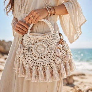 Può includere: Una borsa all'uncinetto color crema caratterizzata da un motivo circolare ispirato ai mandala sul davanti, un unico manico superiore e molteplici nappe pendenti lungo il bordo inferiore, realizzata con complessi punti di filato e nodi.
