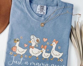 May include: Blue jean Comfort Colors t-shirt with a whimsical illustration of four geese, each adorned with accessories like sunglasses and bows. The phrase "Just a mama goose" is written in cursive below the geese. A gold necklace rests on the shirt.