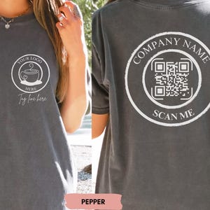May include: A dark grey t-shirt with a white design on the front and back. The front design features a coffee cup with the text "Your Logo Here" and "Tag line here". The back design features a circle with the text "Company Name" and a QR code with the text "Scan Me".