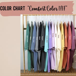 May include: Color chart showing 14 different colors of Comfort Color 1717 t-shirts hanging on a rack. The colors are: Light Green, Moss, Blue Jean, Gray, Black, Pepper, Chalky Mint, Violet, Orchid, White, Ivory, Mustard, Yam, Cumin, and Berry.