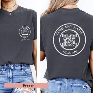 May include: A dark gray t-shirt with a white circle on the front that says "YOUR LOGO HERE" and a coffee cup with steam coming out of it. The back of the shirt has a white circle with the text "COMPANY NAME" and a QR code with the text "SCAN ME" underneath. The shirt is being modeled by a person wearing blue jeans.
