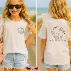 May include: A natural-colored t-shirt with a circular graphic on the front that says "Johnson Family Trip 2023". The back of the shirt has a design that says "Most Likely to Eat All the Snacks" with a beach theme.