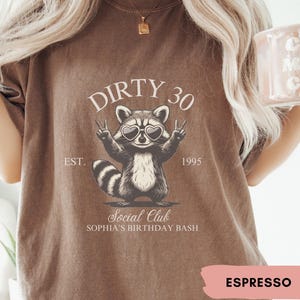 May include: A brown t-shirt with a raccoon wearing sunglasses and the text "Dirty 30 Est. 1995 Social Club Sophia's Birthday Bash" printed on it. The shirt is labeled "Espresso".
