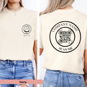 May include: Ivory t-shirt with a black circle on the front featuring a coffee cup with the text "YOUR LOGO HERE" and "Tag line here". The back of the shirt has a black circle with the text "COMPANY NAME" and a QR code with the text "SCAN ME".