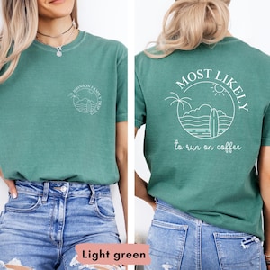May include: Light green t-shirt with a circular graphic on the front and back. The front graphic says "Johnson Family Trip 2023". The back graphic says "MOST LIKELY to run on coffee" with a surfboard, palm trees, and sun design.