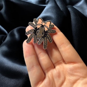 Puede incluir: Un pin de esmalte de araña plateada con un lazo rosa claro. La araña tiene un diseño detallado con ocho patas y un cuerpo gris oscuro. El pin se sostiene en una mano sobre un fondo azul oscuro.
