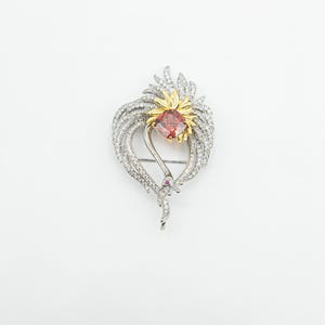 May include: A silver brooch with a heart-shaped design. It features a central, square-cut, reddish-orange gemstone, surrounded by gold accents and clear crystals. A small, pink gemstone is set near the bottom of the brooch.