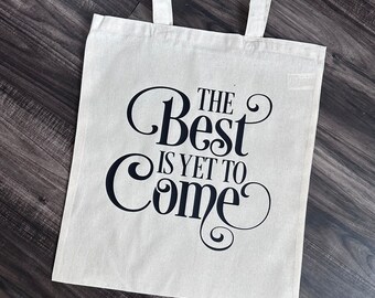 Peut inclure: Un sac fourre-tout en toile blanche avec du texte noir qui dit "The Best is Yet to Come".