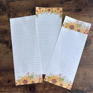 Könnte beinhalten: Drei linierte Notizblöcke mit Sonnenblumen- und Blattmotiven am unteren Rand jeder Seite. Die Notizblöcke sind weiß mit dünnen, hellbraunen Linien.