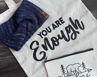 Peut inclure: Un sac fourre-tout crème avec les mots "YOU ARE Enough" en noir. Un bandeau en tricot bleu et violet et une petite pochette zippée crème avec un ours et le mot "explore" sont également visibles.