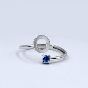 Op de afbeelding: Een zilveren ring met een cirkelvormig ontwerp en een kleine blauwe edelsteen. De ring is aan één kant open en heeft een gebogen band.