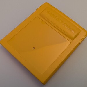 May include: A yellow Game Boy Color cartridge. The cartridge is a rectangular shape with a raised section at the top that reads "GAME BOY Color". The cartridge has a small hole near the center.