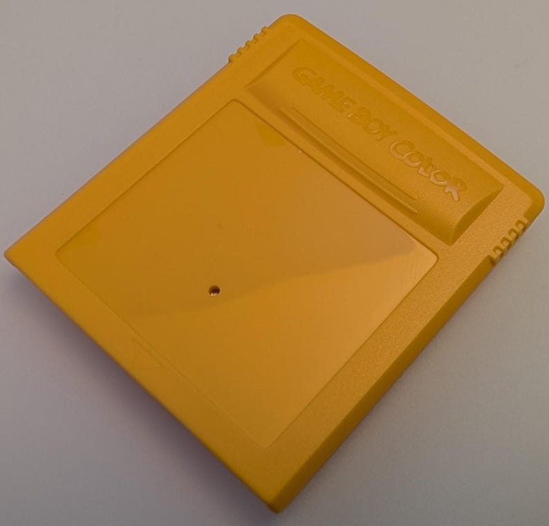 May include: A yellow Game Boy Color cartridge. The cartridge is a rectangular shape with a raised section at the top that reads "GAME BOY Color". The cartridge has a small hole near the center.