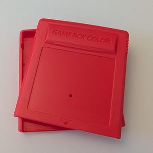 May include: Two bright red Game Boy Color game cartridges are stacked slightly offset. The words "GAME BOY COLOR" are embossed on the top cartridge. The cartridges are made of plastic and have a rectangular shape.