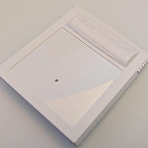 May include: A white Game Boy Color cartridge. The cartridge has a rectangular design with a recessed area and the words "GAME BOY COLOR" embossed on the top. A small, circular indentation is visible on the front.